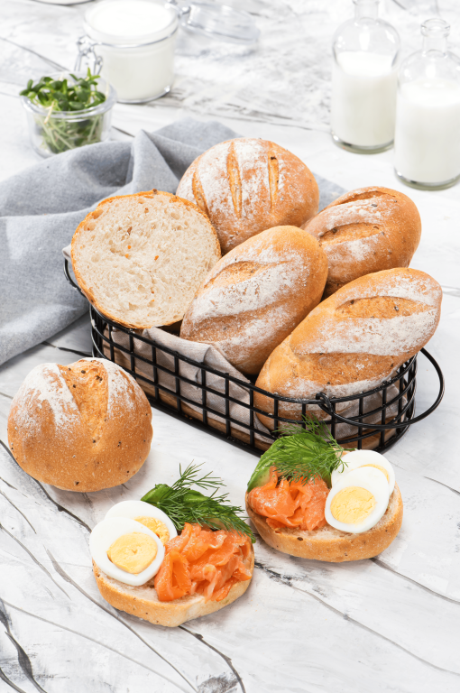 A basket filled with assorted small bread rolls, with slices showing a soft interior, served alongside open sandwiches topped with smoked salmon, boiled eggs, and fresh herbs, with milk bottles in the background