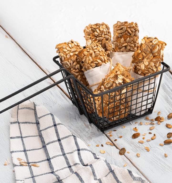 A small basket filled with seeded bread cubes coated in oats and sunflower seeds, placed on a wooden surface with scattered seeds and a folded kitchen towel nearby