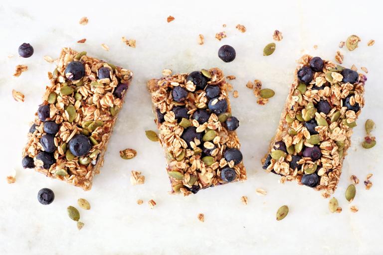 Three rectangular granola bars topped with blueberries, oats, and pumpkin seeds, arranged on a light surface with scattered seeds and crumbs around them